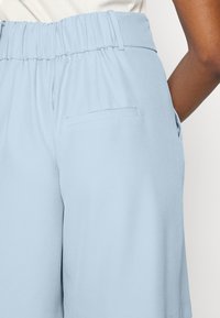 Světle modré šortky s elastickým pasem a zadní kapsou, které nosí osoba s hnědou pletí v bílém topu.