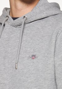Sudadera gris de material de felpa, con capucha ajustable, ojales de metal y un parche de logo en colores rojo, azul y blanco.