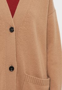 Cardigan beige en maille avec deux boutons noirs et une poche avant, porté sur un vêtement rouge.