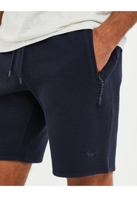 Shorts texturés navy avec un waistband à cordon de serrage, des poches latérales et un détail zippé sur la poche droite.