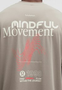Beżowa koszulka sportowa z napisem "Mindful Movement", czerwoną graficzną falą, rokiem "1998" oraz tekstem "Find flow within the journey" na plecach.