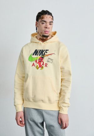 Un sweat à capuche jaune avec un logo graphique Nike noir et rouge, doté d'une poche à l'avant et de poignets côtelés. Porté avec un pantalon de survêtement gris.