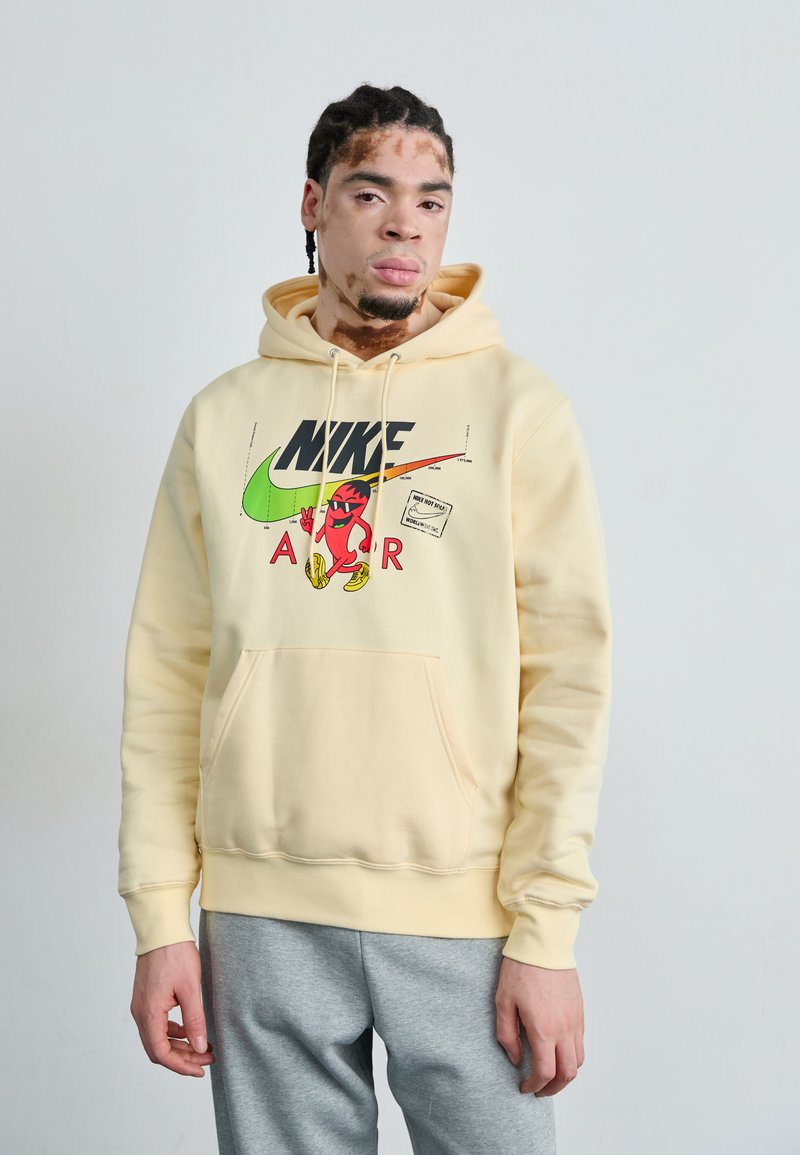 Moletom amarelo com gráfico do logótipo da Nike em preto e vermelho, apresentando um bolso frontal e punhos ribados. Usado com calças de fato cinzentas.