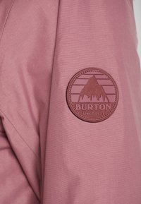 Burtonin logo-patch, jossa on vuoristoteema ja "Since 1977" -teksti, sijaitsee pölyisen vaaleanpunaisen takin hihassa.