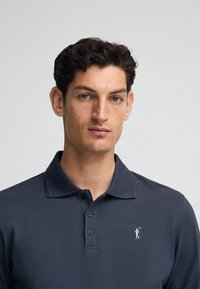 Mörkblå polo skjorta i texturerad tyg, med en treknappsslutning och en liten ljusblå broderad logotyp på vänster bröst.