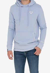 Sudadera azul claro con bolsillo frontal, cordones y un pequeño logo blanco. Material de algodón suave con puños y dobladillo acanalados. Combinada con jeans de mezclilla.