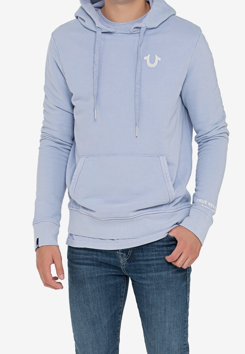 Sudadera azul claro con bolsillo frontal, cordones y un pequeño logo blanco. Material de algodón suave con puños y dobladillo acanalados. Combinada con jeans de mezclilla.