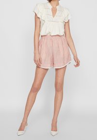 Blouse blanche à volants avec un col en forme de goutte, associée à un short rayé rose clair avec un ourlet en dentelle. Le mannequin porte des talons hauts blancs.