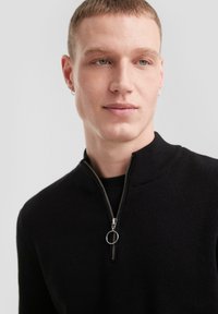 Pull noir zippé avec une texture côtelée, col montant et détail de zip argenté, porté sur une chemise noire. Gros plan du haut du corps.