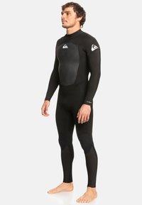 Man staat blote voeten in een zwart wetsuit met lange mouwen, met kniebeschermers en logo's op de borst en de linkermouw.