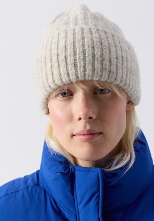 Gros plan sur une personne portant un bonnet en tricot côtelé crème et une doudoune bleue, regardant droit devant avec une expression neutre.