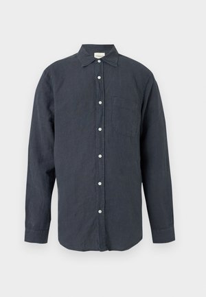 Czarny mężczyzna stojący z ręką w kieszeni, ubrany w ciemną, długą koszulę zapinaną na guziki, niebieskie krótki dżinsowe i czarne skórzane mokasyny.