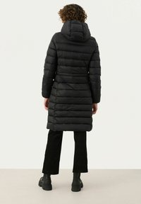 Zwarte pufferjas met een capuchon, voorzien van horizontaal gewatteerde panelen, een getailleerde taille en een jas van gemiddelde lengte. Geklopt met zwarte corduroy broek.