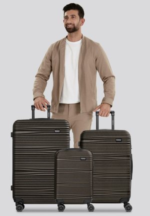 Un homme souriant en tenue beige se tient derrière trois valises texturées marron foncé avec des poignées extensibles sur un fond uni.