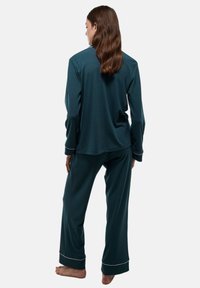 Ensemble de pyjama en teal en tissu doux, avec des manches longues, un pantalon ample et des passepoils blancs au niveau des poignets. Vue de dos, pieds nus.