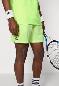 Shorts de sport vert fluo en tissu texturé, avec un logo noir. Associés à un bandeau blanc et tenant une raquette de tennis.