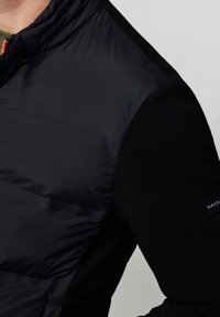 Gros plan sur une personne portant une veste matelassée noire avec des manches en tissu doux et une fermeture éclair partiellement visible près du col.