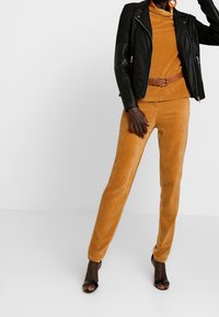 Schwarze Lederbikerjacke über einem senfgelben Cordtop und passenden Hosen; strukturiertes Material; akzentuiert mit einem Gürtel; offene hochhackige Schuhe.