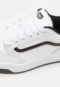 Białe trampki wykonane z materiału syntetycznego, z czarnymi akcentami, płaskimi białymi sznurowadłami, detalami w postaci przeszyć oraz perforowanym wzorem po bokach.