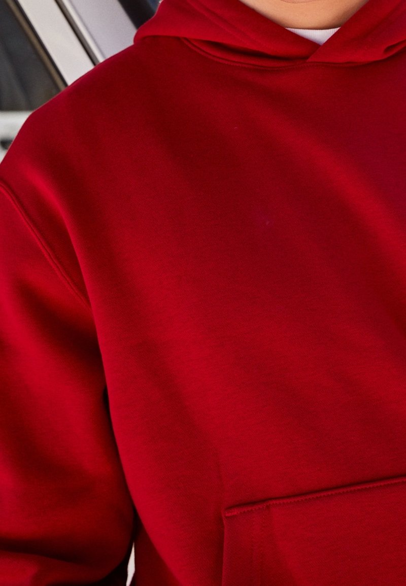 Personne portant un sweat à capuche rouge uni avec une poche avant kangourou, montrant le torse supérieur et une partie du cou.