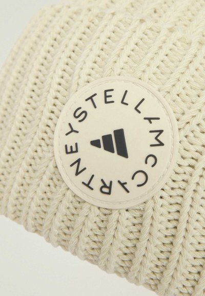 Kreemi värvi kudumine beanie, millel on sooniline tekstuur, sisaldades ümmargust kummitagi musta tekstiga ja kolmnurkse disainiga.