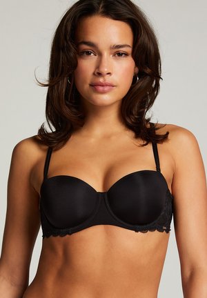 Nő fekete pánt nélküli csipkés szegéllyel ellátott melltartóban, előre nézve, semleges háttér előtt.