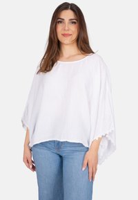 Weißes, strukturiertes Poncho-Top mit lockerer Passform, weitem Ausschnitt und gewelltem Saum. Kombiniert mit blauen Jeans hat es Dreiviertelärmel.