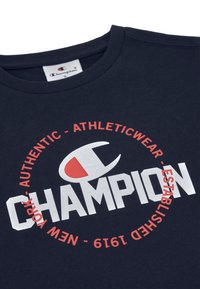 Camiseta de algodón azul marino con el atrevido logo en blanco de "CHAMPION" y texto circular rojo. Diseño de cuello redondo con etiqueta en el cuello.