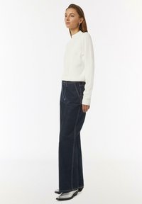 White ribbed long-sleeve top paired with dark blue wide-leg jeans featuring contrasting stitching and black heeled ankle boots.