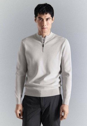 Joven de frente vistiendo un suéter gris claro con cremallera y pantalones negros, contra un fondo gris claro liso.