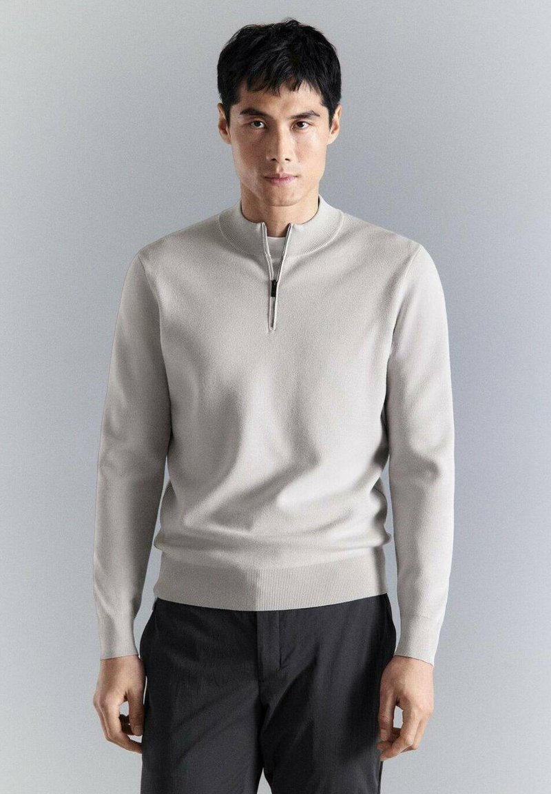 Jeune homme de face portant un pull zippé gris clair et un pantalon noir sur un fond gris clair uni.