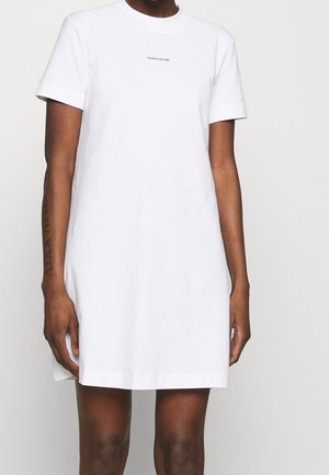 Person wearing a plain white short-sleeve dress with "Calvin Klein Jeans" text on chest, standing against a light background.