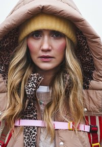 Prešívaná bunda s hnedým vonkajškom, leopardím vzorom na podšívke a ružovými popruhmi. Modelka má na hlave žltú pletenú čiapku a pod bundou nosí svetlý sveter.