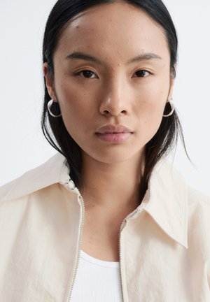 Jeune femme aux cheveux foncés portant des boucles d'oreilles argentées en anneau et une veste beige sur un haut blanc, faisant face à l’avant avec une expression neutre.