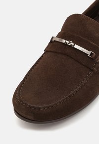 Brown suede loafer with a stitched edge and a metallic Calvin Klein accent across the vamp. Sleek shape with a flat, rounded toe.