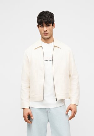 Νεαρός άνδρας που φοράει μπεζ μπουφάν με φερμουάρ πάνω από λευκό T-shirt με γραφικό και ανοιχτό μπλε τζιν, στέκεται μπροστά σε απλό ανοιχτό φόντο.