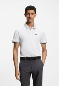 Polo blanco con cuello gris, con un logo negro. Combinado con pantalones gris oscuro y un cinturón negro con hebilla de marca.