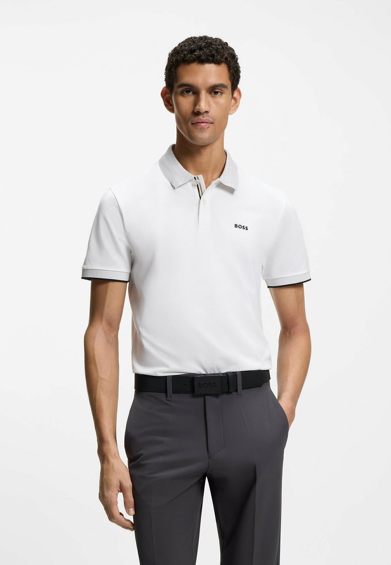 Polo blanco con cuello gris, con un logo negro. Combinado con pantalones gris oscuro y un cinturón negro con hebilla de marca.