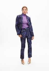 Lila gemusterter Anzug mit Tierdruck, bestehend aus einem taillierten Blazer und maßgeschneiderten Hosen. Kombiniert mit einem lila Rollkragenpullover und goldenen Absatzschuhen.