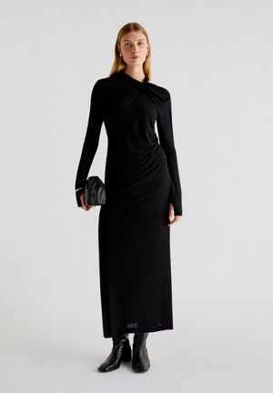 Robe noire à manches longues avec un design drapé et un col noué. Assortie à une pochette noire et des bottes noires jusqu'aux genoux. Texture lisse.