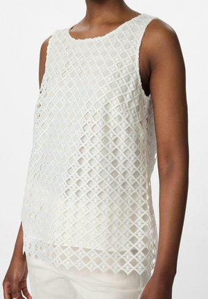 Femme portant un haut blanc sans manches avec un overlay en treillis diamant et un pantalon blanc sur fond blanc uni.