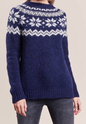 Marineblauer gestrickter Pullover mit weißen Schneeflocken- und Zickzackmustern über Brust und Schultern, kombiniert mit dunkelgrauen Jeans.