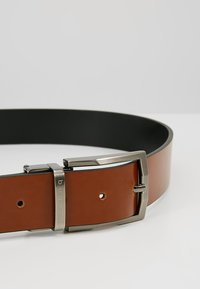 Ceinture en cuir brun avec une doublure intérieure noire et une boucle rectangulaire en métal argenté, comportant un logo et plusieurs trous d'ajustement.