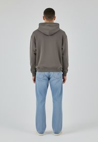 Inimene seisab seljaga kaamera poole, kandes halli kapuutsiga pusa, helesiniseid teksapükse ja valgeid kingi, ühetaolise hele tausta ees.