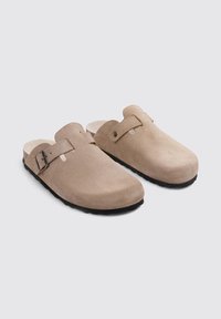 Sabots en suède beige avec une pointe arrondie et une sangle réglable. Semelle en caoutchouc noir et doublure intérieure douce pour le confort.