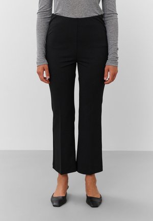 Femme portant un pantalon noir droit, un haut gris à manches longues et des ballerines noires à bouts pointus, debout devant un fond uni.