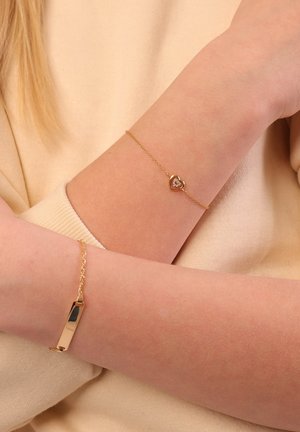 Gouden armbanden om armen; de ene heeft een hart met een centrale steen, de andere is een rechthoekige schakel. Beide hebben een glanzende afwerking.