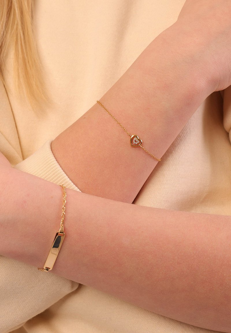 Gouden armbanden om armen; de ene heeft een hart met een centrale steen, de andere is een rechthoekige schakel. Beide hebben een glanzende afwerking.