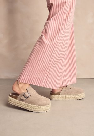 Osoba nosząca beżowe zamszowe buty na platformie typu slip-on z klamrą oraz szerokie spodnie w różowo-białe paski na neutralnej podłodze.