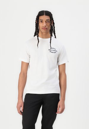 Weißes Baumwoll-T-Shirt mit kurzen Ärmeln, versehen mit einem schwarzen Textlogo auf der linken Brust. Tragen mit schwarzen Hosen. Glatte Textur.